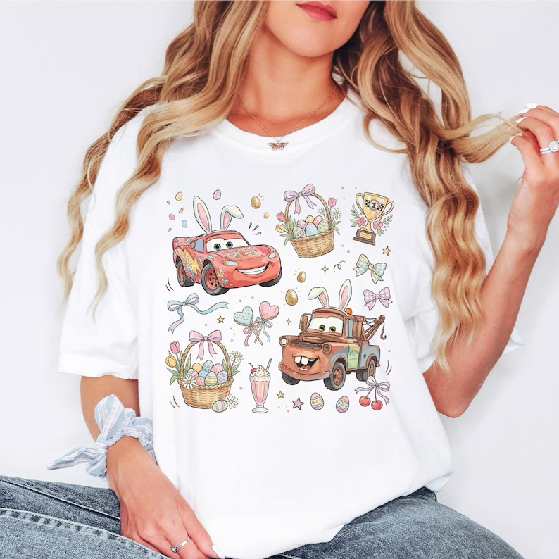 Może przedstawiać: Biała koszulka z kolorową grafiką o tematyce wielkanocnej. Projekt zawiera kresk&oacute;wkowe samochody z kr&oacute;liczymi uszami, koszyki wielkanocne, jajka, kokardy i trofeum. Og&oacute;lny motyw jest świąteczny i zabawny.