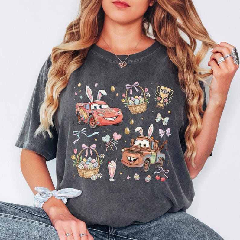 Może przedstawiać: Ciemnoszara koszulka z samochodami z kresk&oacute;wek z kr&oacute;liczymi uszami, koszykami wielkanocnymi, jajkami, kokardkami i trofeum. Projekt zawiera czerwony samoch&oacute;d, brązową lawetę i koktajl mleczny. Koszulka ma wygląd vintage.