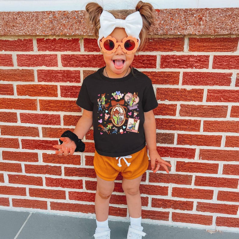 Może przedstawiać: Małe dziecko w czarnym t-shircie z kolorowym nadrukiem graficznym, w połączeniu z musztardowymi szortami. Dziecko ma r&oacute;wnież pomarańczowe okulary przeciwsłoneczne, białą kokardę i białe skarpetki z białymi butami. Tło to czerwona ceglana ściana.