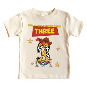 May include: Cream-colored toddler t-shirt with a graphic design. The design features the number "3" in a cowboy theme, with the text "YOU'VE GOT A FRIEND WHOS THREE". Includes cartoon characters and star accents.