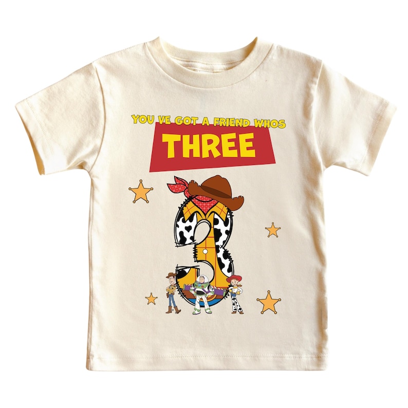 May include: Cream-colored toddler t-shirt with a graphic design. The design features the number "3" in a cowboy theme, with the text "YOU'VE GOT A FRIEND WHOS THREE". Includes cartoon characters and star accents.