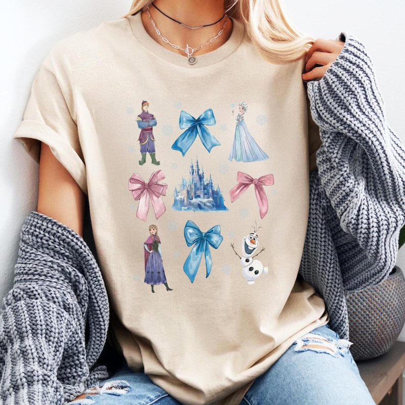 May include: Beige t-shirt featuring a colorful graphic of characters and bows from a popular animated movie. The design includes a castle, snowflakes, and various characters in shades of blue, pink, and purple. The shirt is made of a soft material.