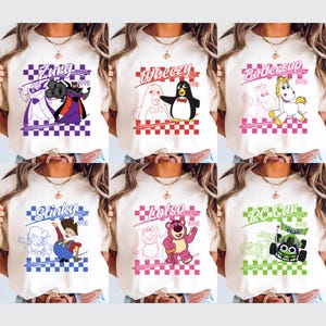 Op de afbeelding: Zes cr&egrave;mekleurige t-shirts met kleurrijke grafische ontwerpen. Elk shirt heeft een ander personage uit een populaire animatiefilm, met tekst boven en onder het personage. De ontwerpen staan tegen een geblokte achtergrond.
