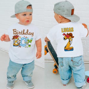 May include: White t-shirt with a colorful "Birthday Boy" graphic featuring a cowboy hat, Woody, Buzz Lightyear, and a rocket ship. The back of the shirt has the name "LUCAS" and a number "2" with a cowboy hat design. The child is wearing light blue jeans and a gray baseball cap.