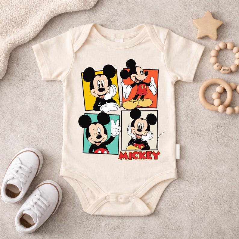 May include: Cream-colored baby onesie featuring four panels with Mickey Mouse illustrations in various poses. The word "MICKEY" is printed in red. White baby shoes and wooden toys are also visible.