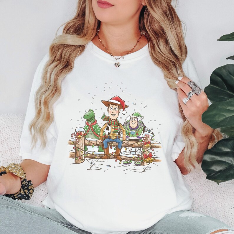 Puede incluir: Camiseta blanca con una ilustraci&oacute;n festiva de Woody, Rex y Buzz Lightyear en una escena nevada. Woody lleva un gorro de Pap&aacute; Noel. El dise&ntilde;o incluye una valla de madera y detalles de bastones de caramelo, evocando un tema navide&ntilde;o.