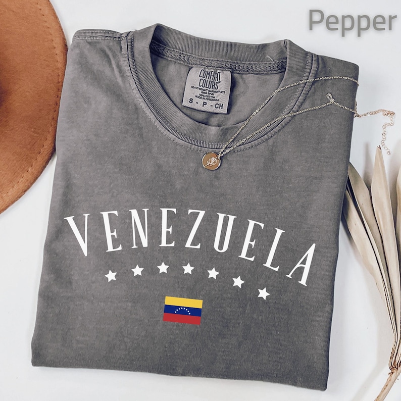 May include: A gray t-shirt with the word "VENEZUELA" in white, arched above a row of white stars and the Venezuelan flag. A silver necklace with a pendant rests on the shirt. The shirt is labeled "Comfort Colors".