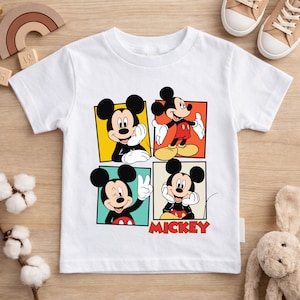 May include: White t-shirt featuring four panels with different poses of Mickey Mouse. Each panel has a different colored background. The word "Mickey" is printed in red at the bottom. The shirt is made of a soft material.