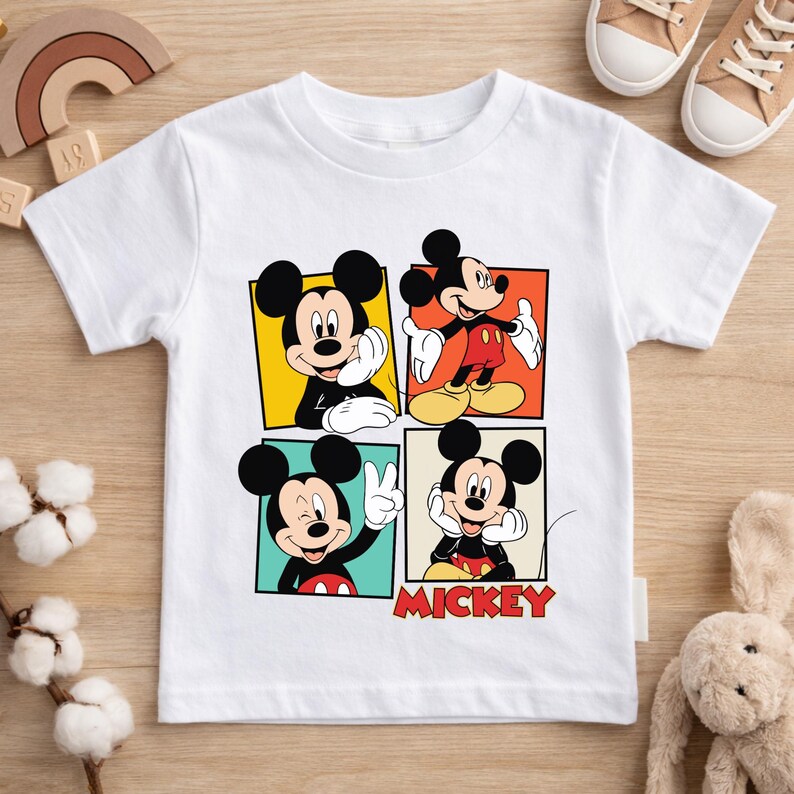 May include: White t-shirt featuring four panels with different poses of Mickey Mouse. Each panel has a different colored background. The word "Mickey" is printed in red at the bottom. The shirt is made of a soft material.