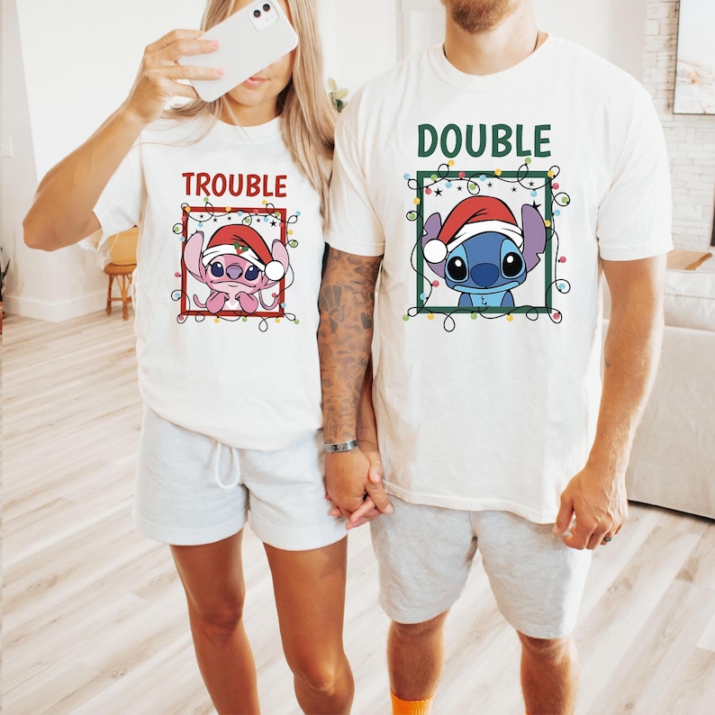May include: Two white t-shirts with matching graphics. One shirt says "TROUBLE" and features a pink cartoon character wearing a Santa hat. The other says "DOUBLE" and has a blue cartoon character in a Santa hat.