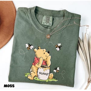 May include: Moss green t-shirt featuring a Winnie the Pooh graphic with bees and a honey pot that says "Hunny". The shirt has a crew neck and short sleeves. A gold necklace is draped across the shirt.