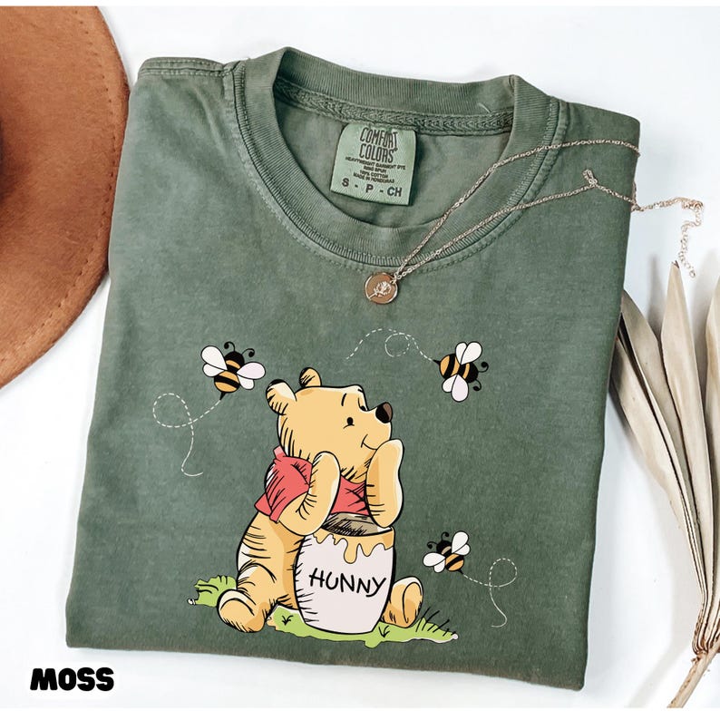 May include: Moss green t-shirt featuring a Winnie the Pooh graphic with bees and a honey pot that says "Hunny". The shirt has a crew neck and short sleeves. A gold necklace is draped across the shirt.