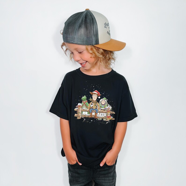 Puede incluir: Un ni&ntilde;o con una camiseta negra con un gr&aacute;fico navide&ntilde;o de los personajes de Toy Story. El ni&ntilde;o tambi&eacute;n lleva una gorra de camionero con la parte trasera de malla gris y la visera marr&oacute;n.