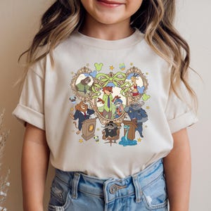May include: A beige t-shirt featuring a colorful cartoon illustration of characters from a popular animated movie. The design includes various animal characters within a decorative frame, with stars and other embellishments.