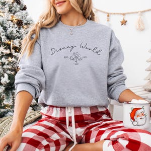 May include: A gray sweatshirt with the words "Disney World" in script, along with an image of Mickey Mouse and the year "1971." The person is wearing red and white plaid pajama pants and holding a mug with a Santa Claus design.