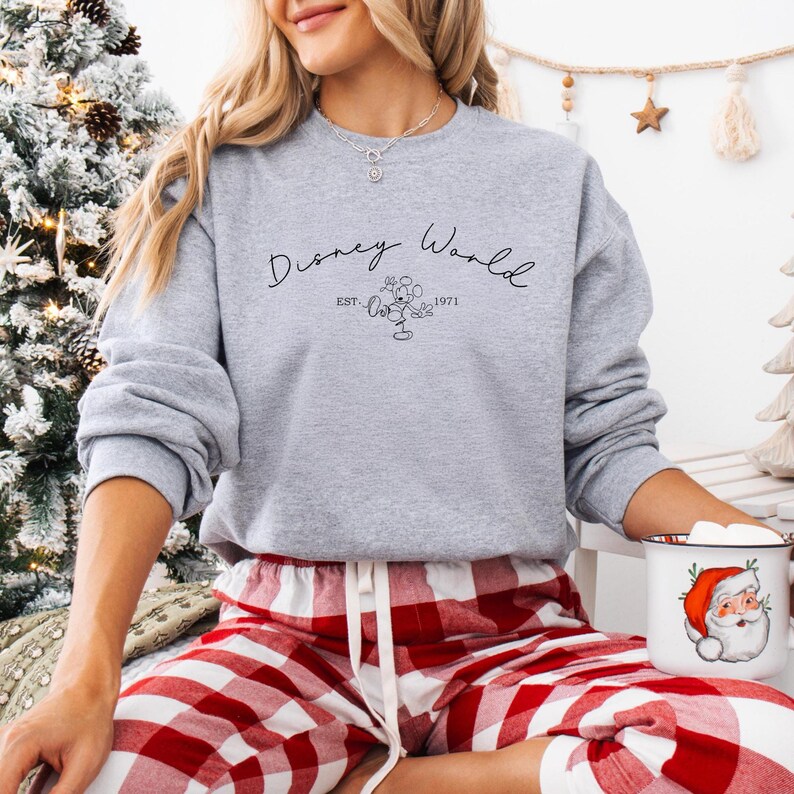 May include: A gray sweatshirt with the words "Disney World" in script, along with an image of Mickey Mouse and the year "1971." The person is wearing red and white plaid pajama pants and holding a mug with a Santa Claus design.