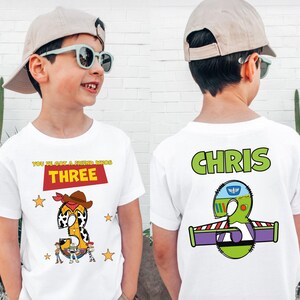May include: White t-shirt with a "Toy Story" theme. The front features the word "THREE" in a red rectangle, a cowboy-themed number 3, and character illustrations. The back displays the name "CHRIS" and a Buzz Lightyear-themed number 3.