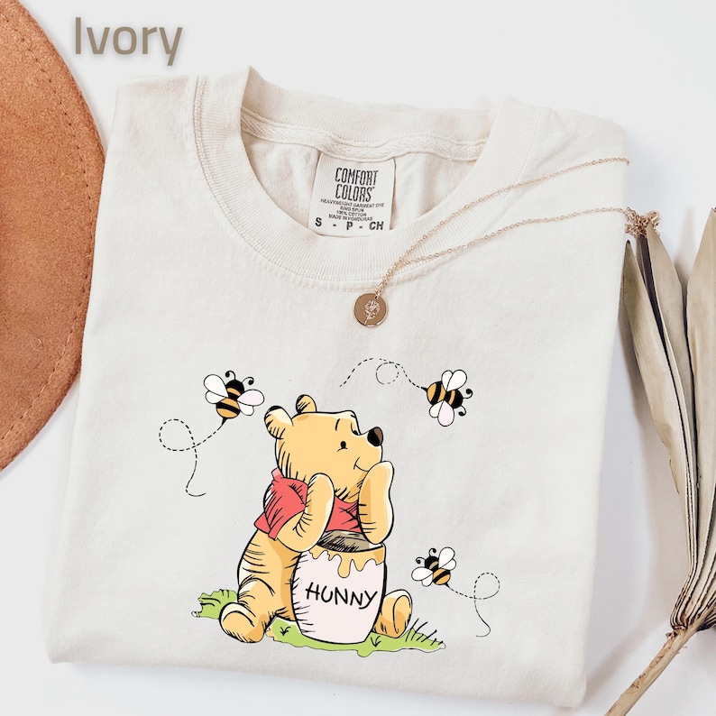May include: Ivory-colored t-shirt featuring a cartoon illustration of Winnie the Pooh with a honey pot labeled "Hunny" and bees. The shirt has a crew neck and short sleeves. A gold necklace with a pendant is also visible.