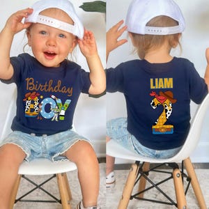 May include: Navy blue t-shirt with "Birthday Boy" text and Toy Story graphics. The back of the shirt features the name "LIAM" and a number 2 design. The child is wearing a white baseball cap and denim shorts.