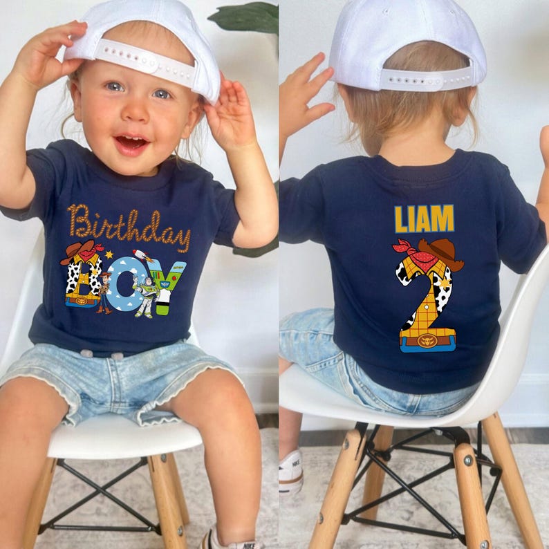 May include: Navy blue t-shirt with "Birthday Boy" text and Toy Story graphics. The back of the shirt features the name "LIAM" and a number 2 design. The child is wearing a white baseball cap and denim shorts.