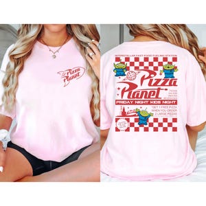 May include: A light pink t-shirt featuring the Pizza Planet logo on the front and back. The back of the shirt has a red and white checkered pattern with the words "Pizza Planet" and "Friday Night Kids Night".
