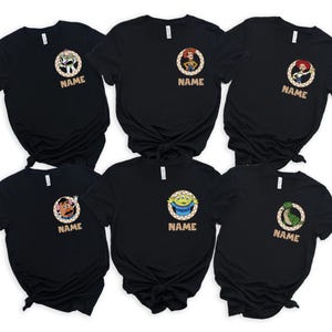 May include: Six black t-shirts featuring characters from a popular animated movie. Each shirt has a circular design with a character image and the word "NAME" below. The characters include Buzz Lightyear, Woody, Jessie, Mr. Potato Head, an alien, and Rex.