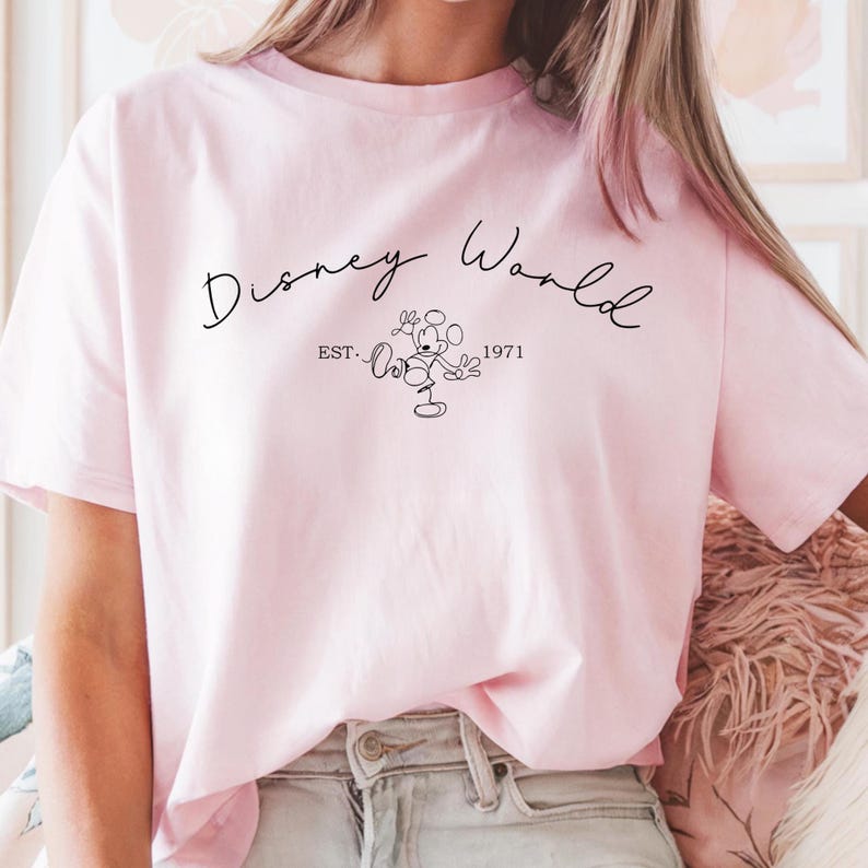 May include: A light pink t-shirt featuring the words "Disney World" in a script font. Below the text is a line drawing of Mickey Mouse and the text "EST. 1971". The shirt is a solid color and appears to be made of a soft material.