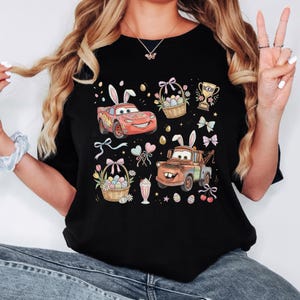 Może przedstawiać: Czarny t-shirt z kolorową grafiką wielkanocną. Projekt zawiera kresk&oacute;wkowe samochody z kr&oacute;liczymi uszami, koszyki wielkanocne wypełnione jajkami, trofeum, kokardy i inne świąteczne elementy. Koszulka to element odzieży codziennej.