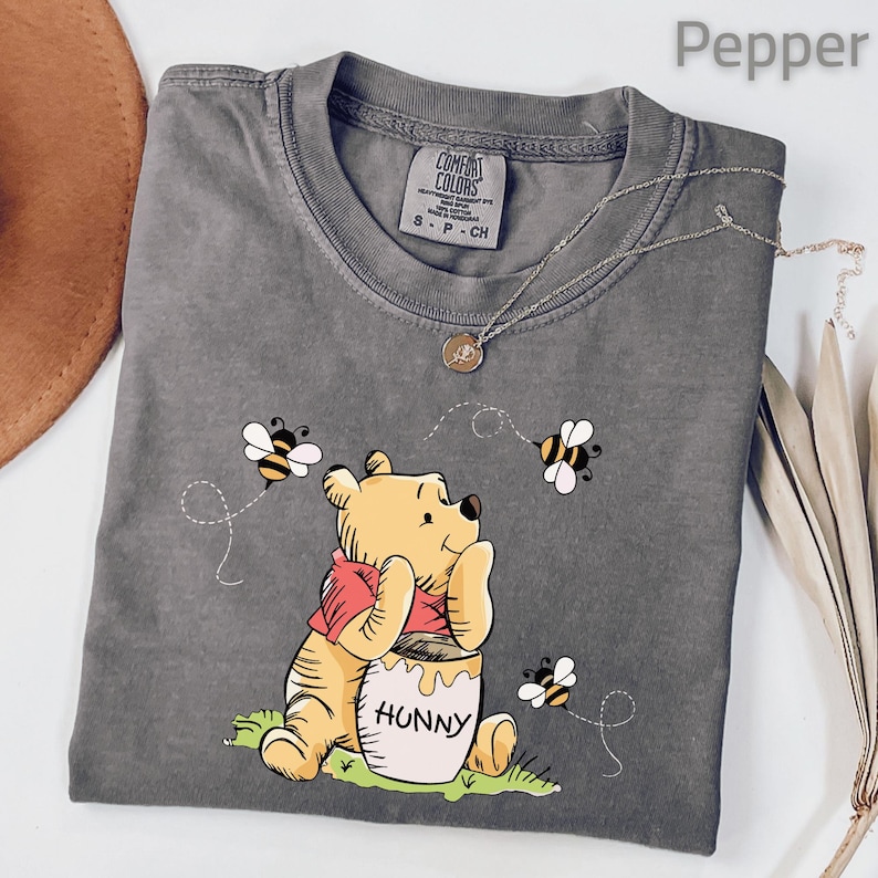 May include: A gray Comfort Colors t-shirt featuring a Winnie the Pooh graphic. The cartoon character is holding a honey pot with the word "Hunny" on it, surrounded by bees. A gold necklace is draped across the shirt.