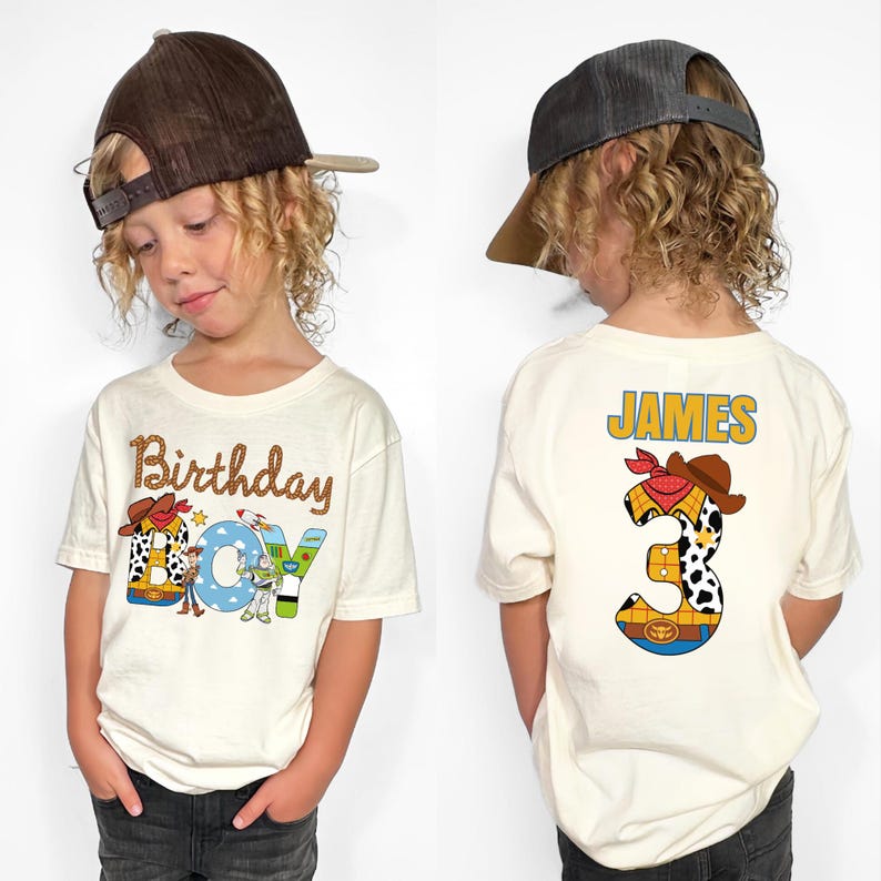 May include: Cream-colored t-shirt with a colorful "Birthday BOY" graphic featuring a cowboy hat and Toy Story characters. The back of the shirt displays the name "JAMES" and a number "3" with a cowboy theme. The child is wearing a brown baseball cap.