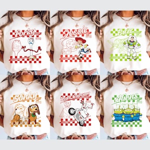 Op de afbeelding: Cr&egrave;mekleurige T-shirts met verschillende Toy Story-personages. Elk shirt heeft een personagenaam in een gestileerd lettertype, een afbeelding en het jaar 1995. Personages zijn Forky, Jessie, Buzz, Slinky, Duke Caboom en Aliens.