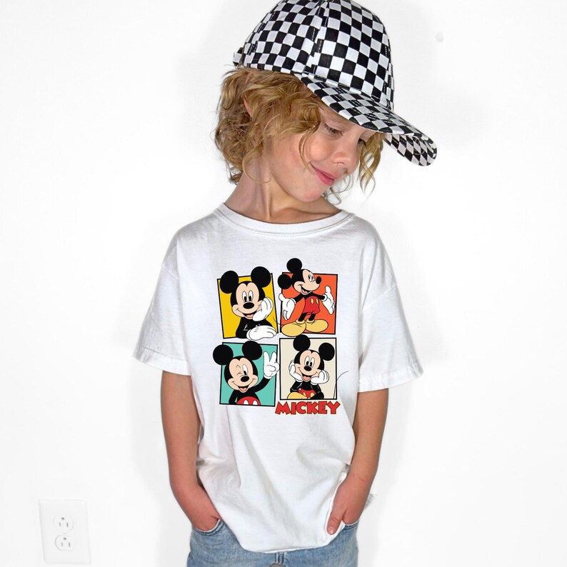 May include: A white t-shirt featuring four panels with different images of Mickey Mouse. The word "Mickey" is printed in red. The person is wearing a black and white checkered baseball cap.