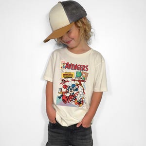 May include: A child wearing a cream-colored t-shirt featuring a comic book-style graphic with the word "Avengers" in red. The shirt showcases Disney characters like Mickey Mouse and Donald Duck. The child is also wearing a baseball cap.