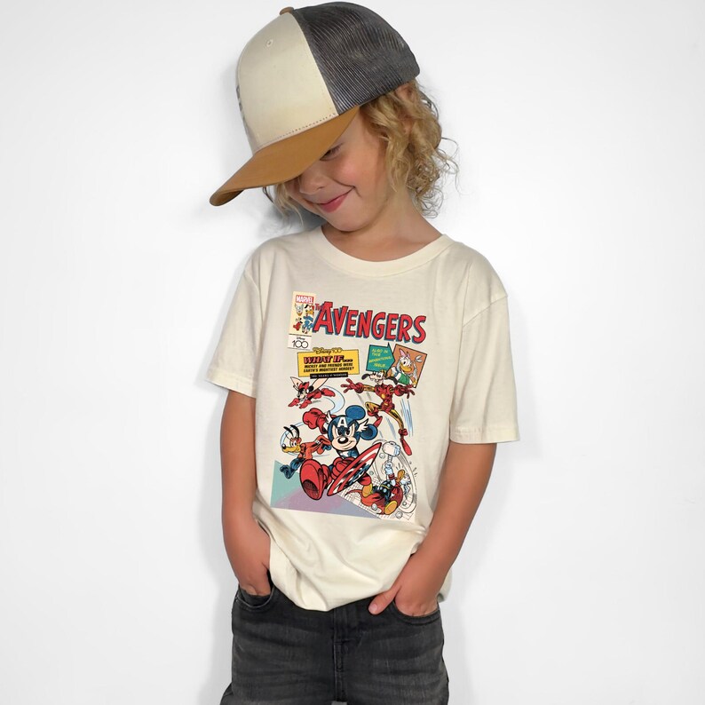 May include: A child wearing a cream-colored t-shirt featuring a comic book-style graphic with the word "Avengers" in red. The shirt showcases Disney characters like Mickey Mouse and Donald Duck. The child is also wearing a baseball cap.