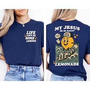 Puede incluir: Camiseta azul marino con el texto "LIFE GAVE ME SOME LEMONS" en la parte delantera. La parte trasera presenta un lim&oacute;n de dibujos animados surfeando con el texto "MY JESUS HE BE MAKIN'" y "LEMONADE".