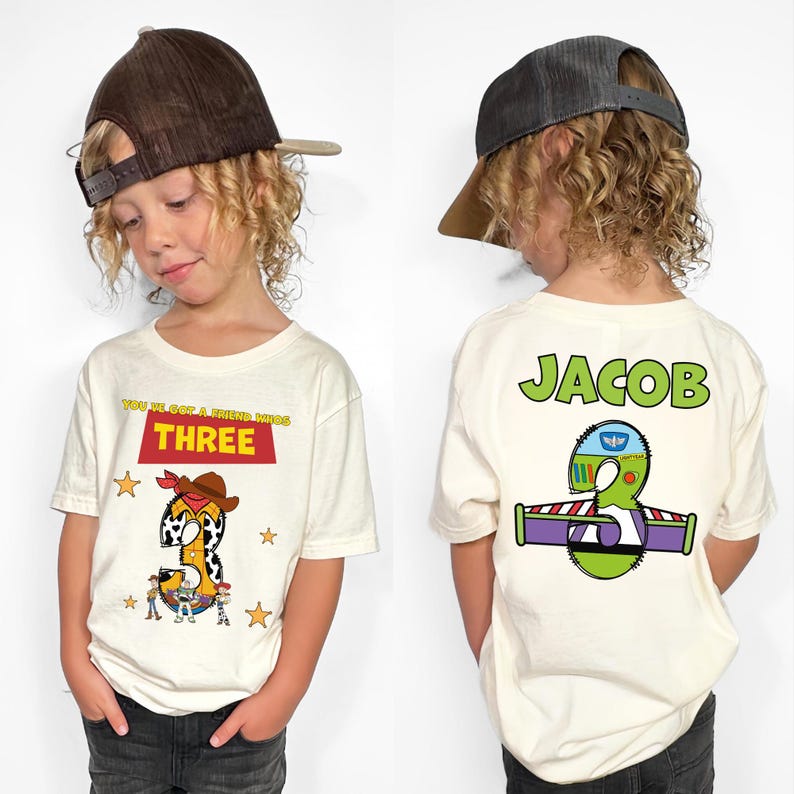 May include: Off-white short-sleeved t-shirt with a "Toy Story" theme. The front features a "THREE" graphic with a cowboy-themed number 3. The back displays the name "JACOB" and a Buzz Lightyear-themed number 3.