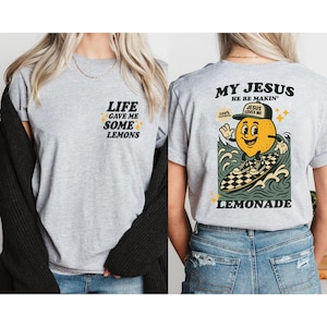 Puede incluir: Camiseta gris claro con el texto "LIFE GAVE ME SOME LEMONS" en la parte delantera. La parte trasera de la camiseta presenta un lim&oacute;n de dibujos animados surfeando con el texto "MY JESUS HE BE MAKIN' LEMONADE".