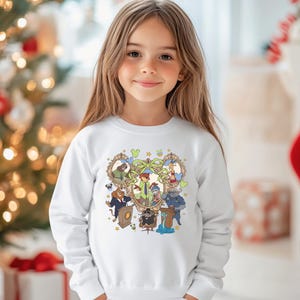 May include: A white sweatshirt featuring a colorful cartoon graphic. The graphic includes several animal characters in ornate frames, with a central character in a green suit. The design is surrounded by decorative elements.
