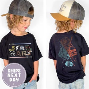 May include: Black Star Wars t-shirt featuring the Star Wars logo and characters on the front and back. The front design includes the Star Wars logo with lightsabers. The back design features Darth Vader, a Stormtrooper, and other characters.