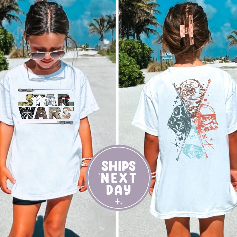 May include: White t-shirt featuring the Star Wars logo on the front and character graphics on the back. The front design includes the words "STAR WARS" in a colorful, blocky font with lightsabers. The back displays Darth Vader, a Stormtrooper, and the Death Star.