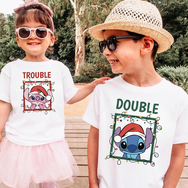 May include: Two children wearing white t-shirts with cartoon character designs. One shirt says "TROUBLE" and features a pink alien in a Santa hat. The other shirt says "DOUBLE" and features a blue alien in a Santa hat. Both designs are framed with Christmas lights.