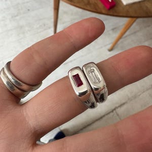 K&ouml;nnte beinhalten: Nahaufnahme von zwei silbernen Ringen mit rechteckigen Edelsteinen, einem roten und einem klaren. Links ist ein Stapel von drei silbernen Ringen zu sehen. Die Ringe werden auf dem Finger einer Person pr&auml;sentiert.