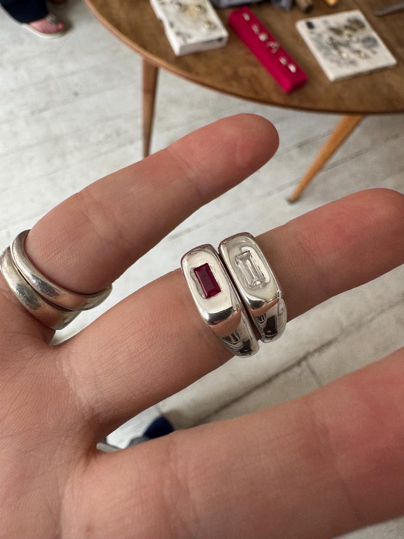 K&ouml;nnte beinhalten: Nahaufnahme von zwei silbernen Ringen mit rechteckigen Edelsteinen, einem roten und einem klaren. Links ist ein Stapel von drei silbernen Ringen zu sehen. Die Ringe werden auf dem Finger einer Person pr&auml;sentiert.