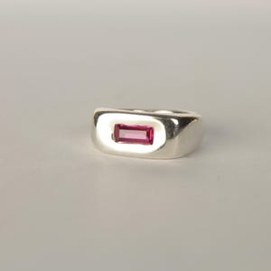 K&ouml;nnte beinhalten: Ein silberner Ring mit einem rechteckigen, magentafarbenen Edelstein. Der Ring hat eine polierte Oberfl&auml;che und ein dickes Band. Der Edelstein ist zentral gesetzt und bildet einen auff&auml;lligen Kontrast.
