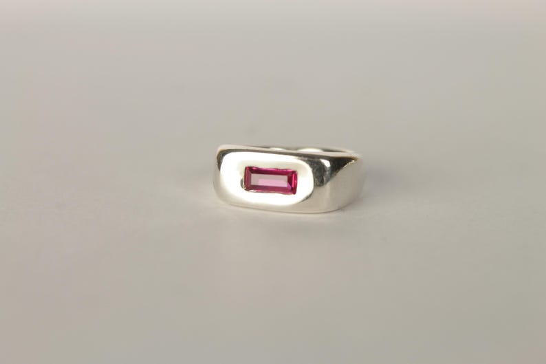 K&ouml;nnte beinhalten: Ein silberner Ring mit einem rechteckigen, magentafarbenen Edelstein. Der Ring hat eine polierte Oberfl&auml;che und ein dickes Band. Der Edelstein ist zentral gesetzt und bildet einen auff&auml;lligen Kontrast.
