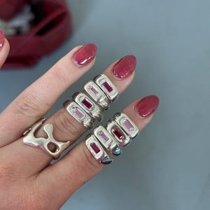 K&ouml;nnte beinhalten: Eine Hand zeigt mehrere silberne Ringe mit rechteckigen, rosa Edelsteinen. Ein Ring hat eine organische Form. Die Ringe werden an mehreren Fingern getragen und pr&auml;sentieren das Design und die Farbe des Schmucks vor neutralem Hintergrund.