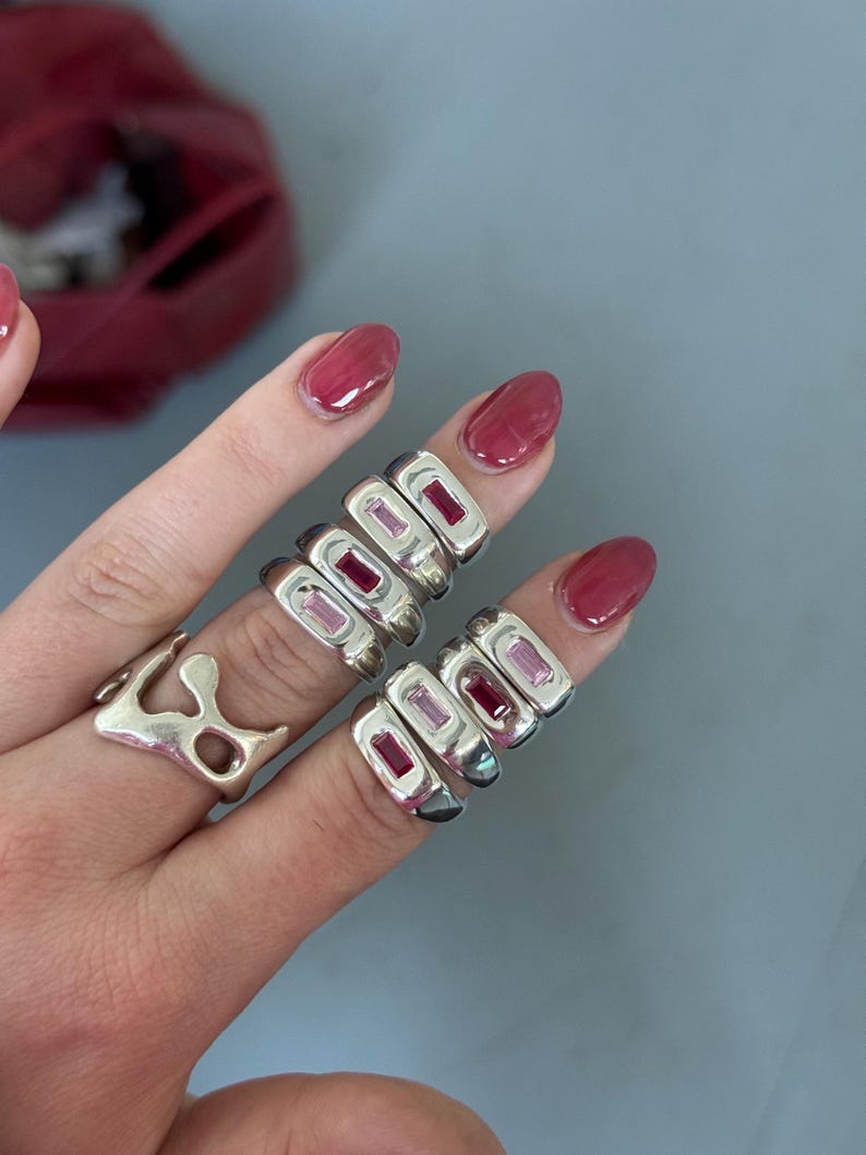 K&ouml;nnte beinhalten: Eine Hand zeigt mehrere silberne Ringe mit rechteckigen, rosa Edelsteinen. Ein Ring hat eine organische Form. Die Ringe werden an mehreren Fingern getragen und pr&auml;sentieren das Design und die Farbe des Schmucks vor neutralem Hintergrund.