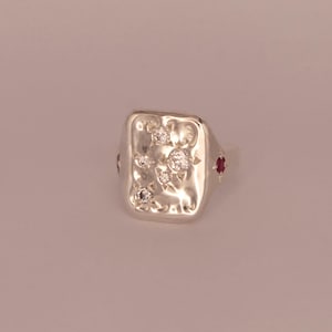May include: A silver signet ring with a rectangular face. The face is textured and decorated with several small, clear gemstones. Two small red gemstones are set on the sides of the ring.