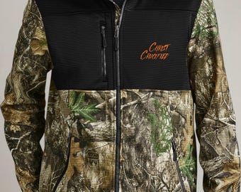 May include: A full-zip jacket with a black upper section and a camouflage lower section. The jacket features the text "Christ Crucified" in orange script. It has two zippered pockets and a ribbed texture on the black portion.