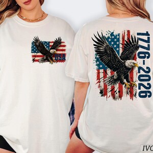 Pode incluir: Uma t-shirt cor de marfim com o gr&aacute;fico de uma &aacute;guia-de-cabe&ccedil;a-branca a voar sobre uma bandeira americana desgastada. O peito apresenta uma pequena &aacute;guia e a bandeira com o texto USA. As costas mostram uma grande &aacute;guia, a bandeira e o texto 1776-2026 em azul.